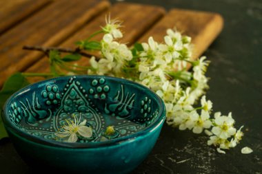 Ahşap bir tahtada beyaz kiraz çiçeklerinin arka planında şeffaf su bulunan mavi bir Türk kasesi ve siyah bir arka plan. Uzayı kopyala Bahar tatili kavramı