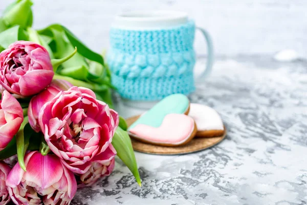 Bouquet of pink tulips, a cup of tea, and sweet gingerbread cookies in the shape of a heart on a light background. Copy space. Spring holiday concept, Valentine\'s Day
