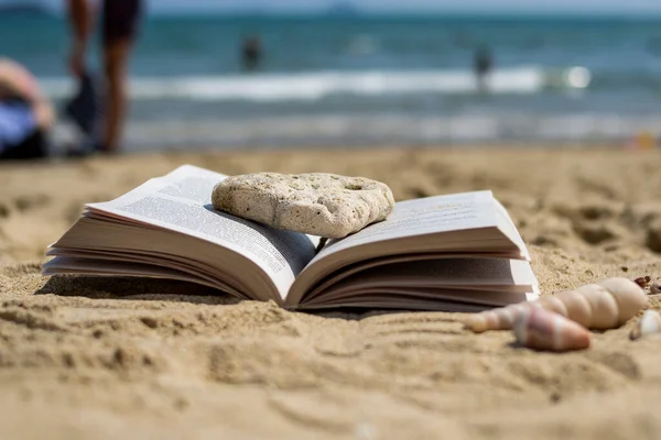 stock image An open book crushed by a stone lies on the sand on the beach. Blurred background. Summer vacation on the beach.