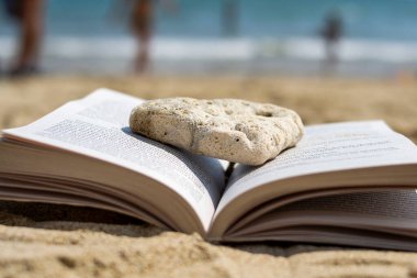Sahilde kumsalda bir taş tarafından ezilmiş açık bir kitap yatıyor. Arka plan bulanık. Plajda yaz tatili.