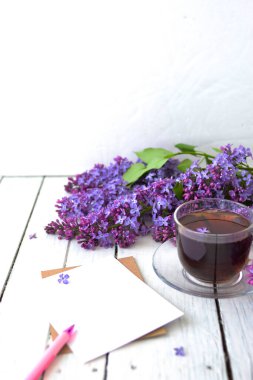 Leylak çiçekleri, şeffaf bir fincan çay ve bir fincan çay, beyaz bir vazo ve notlar için el işi kağıt ve beyaz ahşap bir masanın üzerinde pembe bir kalem. Modelleme. Uzayı kopyala Bahar kahvaltısı kavramı.