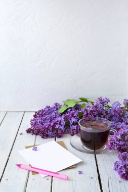 Leylak çiçekleri, şeffaf bir fincan çay ve bir fincan çay, beyaz bir vazo ve notlar için el işi kağıt ve beyaz ahşap bir masanın üzerinde pembe bir kalem. Modelleme. Uzayı kopyala Bahar kahvaltısı kavramı.