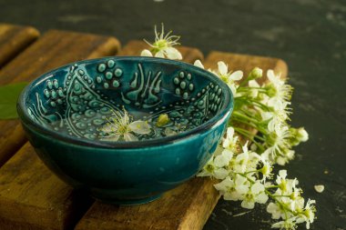Ahşap bir tahtada beyaz kiraz çiçeklerinin arka planında şeffaf su bulunan mavi bir Türk kasesi ve siyah bir arka plan. Uzayı kopyala Bahar tatili kavramı
