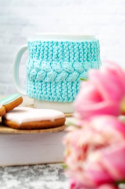 Örülmüş mavi bir çantanın içinde beyaz bir fincan çay bir kitap, pembe çiçekler, laleler ve dokuma masasındaki zencefilli ekmeğin üzerinde duruyor. Bahar konsepti.