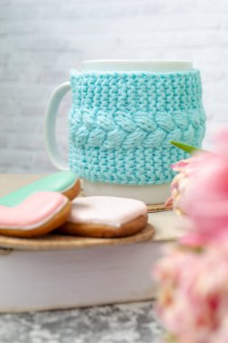 Örülmüş mavi bir çantanın içinde beyaz bir fincan çay bir kitap, pembe çiçekler, laleler ve dokuma masasındaki zencefilli ekmeğin üzerinde duruyor. Bahar konsepti.