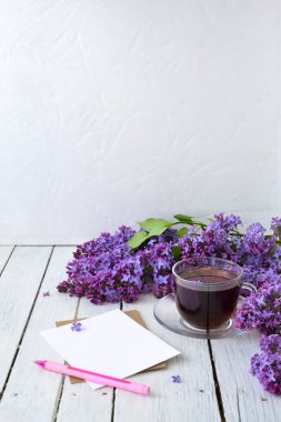 Leylak çiçekleri, şeffaf bir fincan çay ve bir fincan çay, beyaz bir vazo ve notlar için el işi kağıt ve beyaz ahşap bir masanın üzerinde pembe bir kalem. Modelleme. Uzayı kopyala Bahar kahvaltısı kavramı.