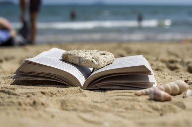 Sahilde kumsalda bir taş tarafından ezilmiş açık bir kitap yatıyor. Arka plan bulanık. Plajda yaz tatili.