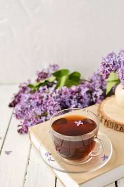 Romantik bir arka plan, bir fincan çay, leylak çiçekleri ve beyaz ahşap bir masanın üzerinde bir kitap. Eğlence konsepti, bahar kahvaltısı.