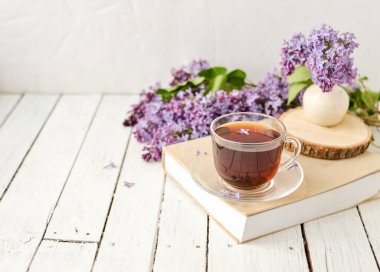 Romantik bir arka plan, bir fincan çay, leylak çiçekleri ve beyaz ahşap bir masanın üzerinde bir kitap. Eğlence konsepti, bahar kahvaltısı.