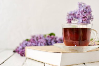 Romantik bir arka plan, bir fincan çay, leylak çiçekleri ve beyaz ahşap bir masanın üzerinde bir kitap. Eğlence konsepti, bahar kahvaltısı..