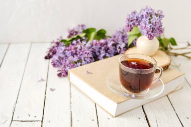 Romantik bir arka plan, bir fincan çay, leylak çiçekleri ve beyaz ahşap bir masanın üzerinde bir kitap. Eğlence konsepti, bahar kahvaltısı..