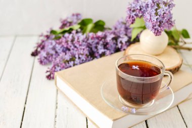 Romantik bir arka plan, bir fincan çay, leylak çiçekleri ve beyaz ahşap bir masanın üzerinde bir kitap. Eğlence konsepti, bahar kahvaltısı..