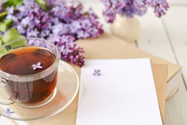 Romantik bir arka plan, bir fincan çay, leylak çiçekleri ve beyaz ahşap bir masanın üzerinde bir kitap. Boş zaman konsepti, bahar kahvaltısı. Model