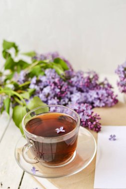 Romantik bir arka plan, bir fincan çay, leylak çiçekleri ve beyaz ahşap bir masanın üzerinde bir kitap. Eğlence konsepti, bahar kahvaltısı..