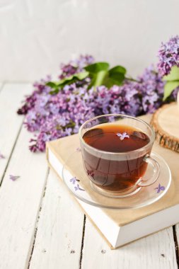 Romantik bir arka plan, bir fincan çay, leylak çiçekleri ve beyaz ahşap bir masanın üzerinde bir kitap. Eğlence konsepti, bahar kahvaltısı..