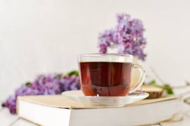 Romantik bir arka plan, bir fincan çay, leylak çiçekleri ve beyaz ahşap bir masanın üzerinde bir kitap. Eğlence konsepti, bahar kahvaltısı.