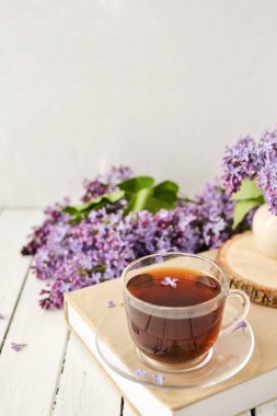 Romantik bir arka plan, bir fincan çay, leylak çiçekleri ve beyaz ahşap bir masanın üzerinde bir kitap. Eğlence konsepti, bahar kahvaltısı.