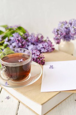 Romantik bir arka plan, bir fincan çay, leylak çiçekleri ve beyaz ahşap bir masanın üzerinde bir kitap. Boş zaman konsepti, bahar kahvaltısı. Model.