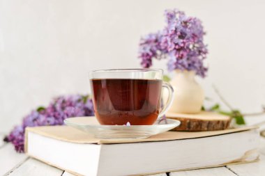 Romantik bir arka plan, bir fincan çay, leylak çiçekleri ve beyaz ahşap bir masanın üzerinde bir kitap. Eğlence konsepti, bahar kahvaltısı.
