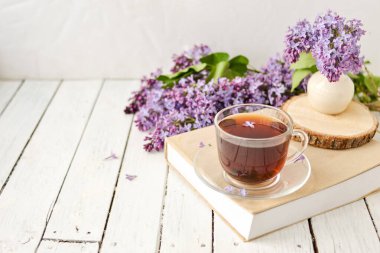 Romantik bir arka plan, bir fincan çay, leylak çiçekleri ve beyaz ahşap bir masanın üzerinde bir kitap. Eğlence konsepti, bahar kahvaltısı.