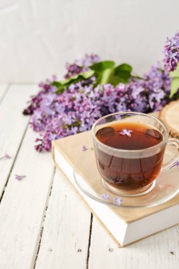 Romantik bir arka plan, bir fincan çay, leylak çiçekleri ve beyaz ahşap bir masanın üzerinde bir kitap. Eğlence konsepti, bahar kahvaltısı.