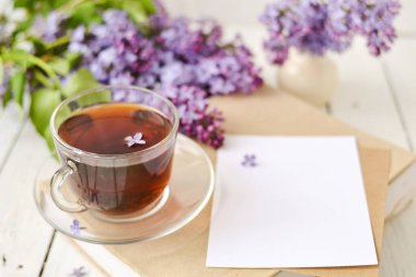 Romantik bir arka plan, bir fincan çay, leylak çiçekleri ve beyaz ahşap bir masanın üzerinde bir kitap. Boş zaman konsepti, bahar kahvaltısı. Model.