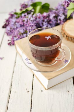 Romantik bir arka plan, bir fincan çay, leylak çiçekleri ve beyaz ahşap bir masanın üzerinde bir kitap. Eğlence konsepti, bahar kahvaltısı.