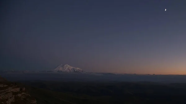 Güneşin son ışınlarında Elbrus koyu mavi bir gökyüzü ve yeni bir ayın arka planına karşı. Manzara Bermamyt Platosu, Karachay-Cêess Cumhuriyeti, Rusya