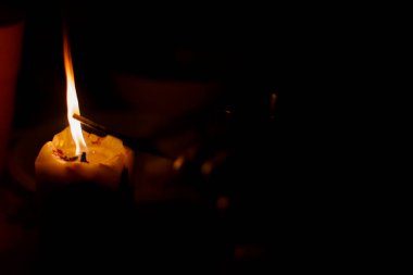 Karanlık bir arka planda bir mum yanıyor. Yanında ince bir sopa var.