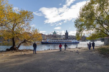 Prag, Çek Cumhuriyeti - 3 Kasım 2025: Vltava Nehri 'nin kumlu kıyısında bir grup insan Prag' ın arka planında tarihi mimariyle nehir kenarındaki manzarayı gözlemliyor.