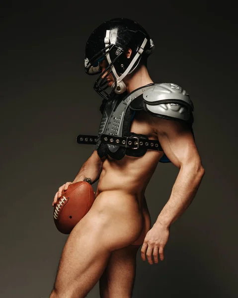 Naked american football player covering with ball at grey background