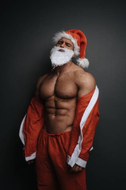 Stüdyoda gri arka planda Noel Baba şapkalı kibirli kaslı adam. Noel görünüşlü yakışıklı bir adam. Kırmızı Noel Baba kostümlü seksi iri kıyım..