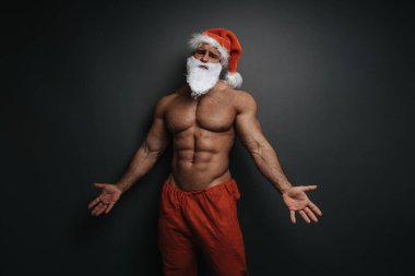 Noel Baba şapkalı gömleksiz kaslı adam ellerini yana doğru tutuyor. Stüdyoda gri arka planda seksi bir Noel erkeği..