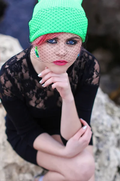 Hermosa adolescente con ojos azules en un sombrero de moda y elegante color verde con velo negro sentado junto al mar en las rocas amarillo — Foto de Stock