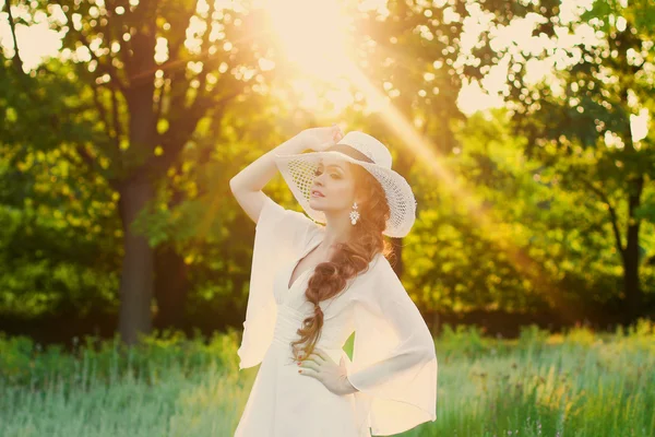 Hermosa chica pelirroja con un elegante sombrero de paja en un jardín botánico en los rayos del sol poniente. Poner el sol directamente detrás de ella crea una luz cálida y suave en la escena. Silueta dorada . — Foto de Stock