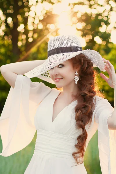 漂亮的红发女孩时尚的草帽，在夕阳的光线的植物园。设置直接在她身后的太阳在场景上创建温暖，光线柔和。金色的剪影. — 图库照片