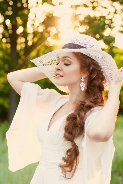 Beautiful red-haired girl in a stylish straw hat in a botanical garden in the rays of the setting sun. Setting sun directly behind her creates a warm and soft light on scene. Golden silhouette. — Stock Photo, Image