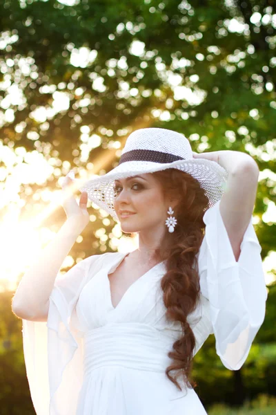 Hermosa chica pelirroja con un elegante sombrero de paja en un jardín botánico en los rayos del sol poniente. Poner el sol directamente detrás de ella crea una luz cálida y suave en la escena. Silueta dorada . —  Fotos de Stock