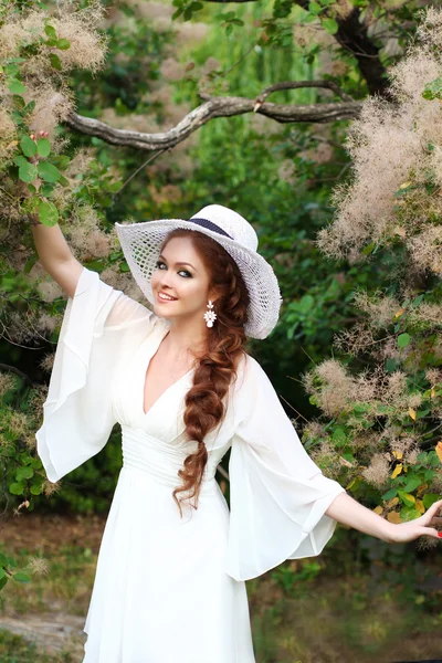 Belle fille aux cheveux roux au chapeau de paille élégant, portant une élégante robe blanche dans un jardin botanique aux rayons du soleil couchant . — Photo