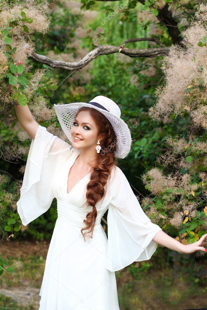 Beautiful red-haired girl in a stylish straw hat, wearing elegant white dress in a botanical garden in the rays of the setting sun.