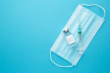 Coronavirus veya COVID 19, 2019 - mavi arka planda şırınga ve hijyen koruyucu yüz maskesi olan bir şişedeki nCoV aşısı. Üst görünüm