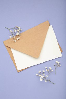 Wedding invitation concept. Craft envelope with paper card and white gypsophila flowers on pastel purple background. Top view