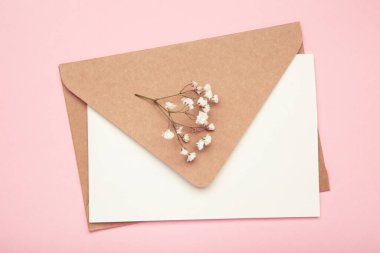 Wedding invitation concept. Craft envelope with paper card and white gypsophila flowers on pastel pink background. Top view