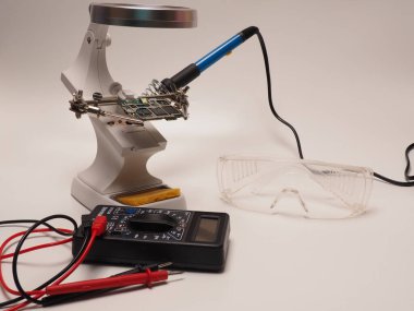Elektronik tamir aletlerinin resmi, PCB tutucu standı, lehimleme demiri, multimetre ve koruyucu gözlüklerden oluşur. İzole edilmiş beyaz bir arkaplanda çekim yap