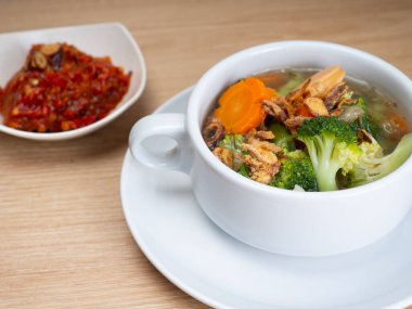 Sosis, brokoli, havuç içeren sebze çorbası beyaz bir porselen kasede küçük bir kase kırmızı biber ezmesi.