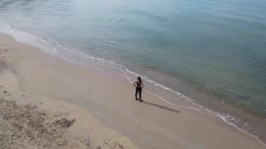 Asyalı kadın koşucunun açık deniz kumsalında ve beyaz kumsalda gün doğumunda plaj boyunca koştuğunu gösteren hava görüntüsü.