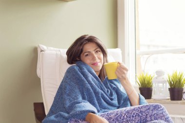 Evde oturan genç kadın, elinde bir fincan çayla pencereden dışarı bakıyor.