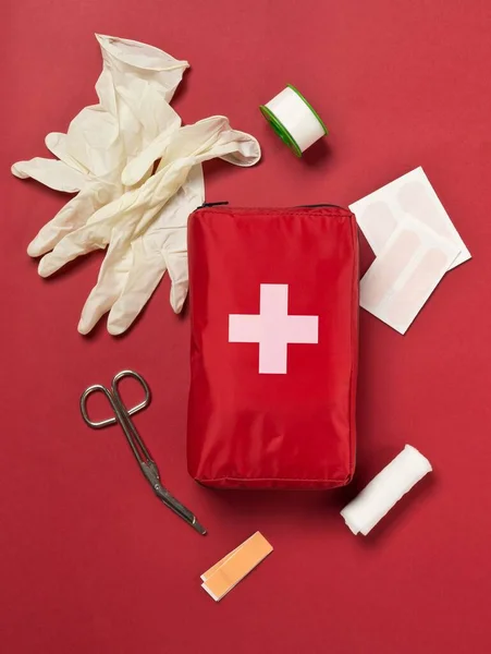 Red first aid medical kit bag with scissors, tape and gloves top view flat lay from above over red background