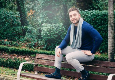 Genç bir adam bankta oturuyor ve kameraya bakıyor.