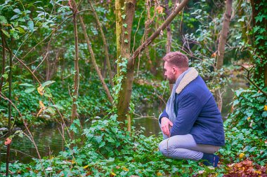 Sonbaharda nehrin kenarındaki ormanda çömelmiş genç bir adam.